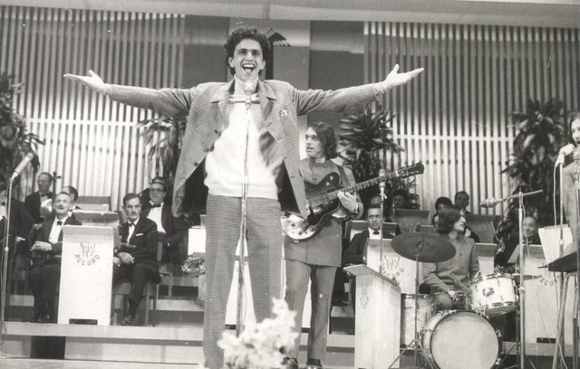 A imagem mostra Caetano Veloso de frente e cantando ao microfone com os braços abertos