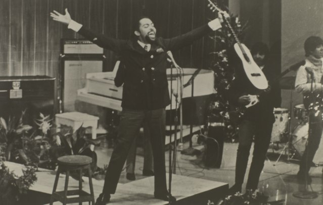 A imagem mostra Gilberto Gil cantando de braços abertos, segurando um violão em uma das mãos, nos anos 1960
